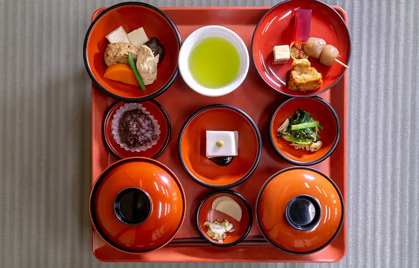 Traditional Japanese shojin-ryori meal beautifully arranged in lacquer bowls on a red tray, featuring colorful seasonal vegetables, tofu, and green tea.