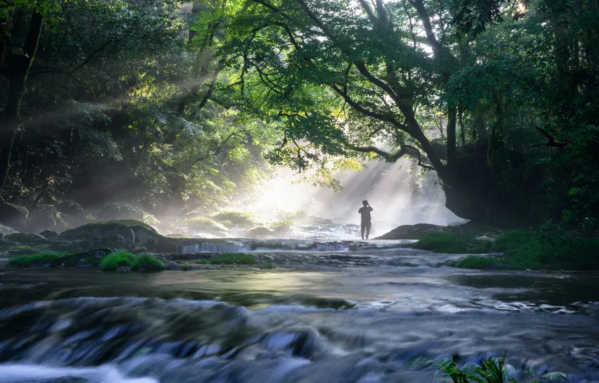 A back-view photo of a person standing by a waterfall with their arms spread wide, inhaling the air.